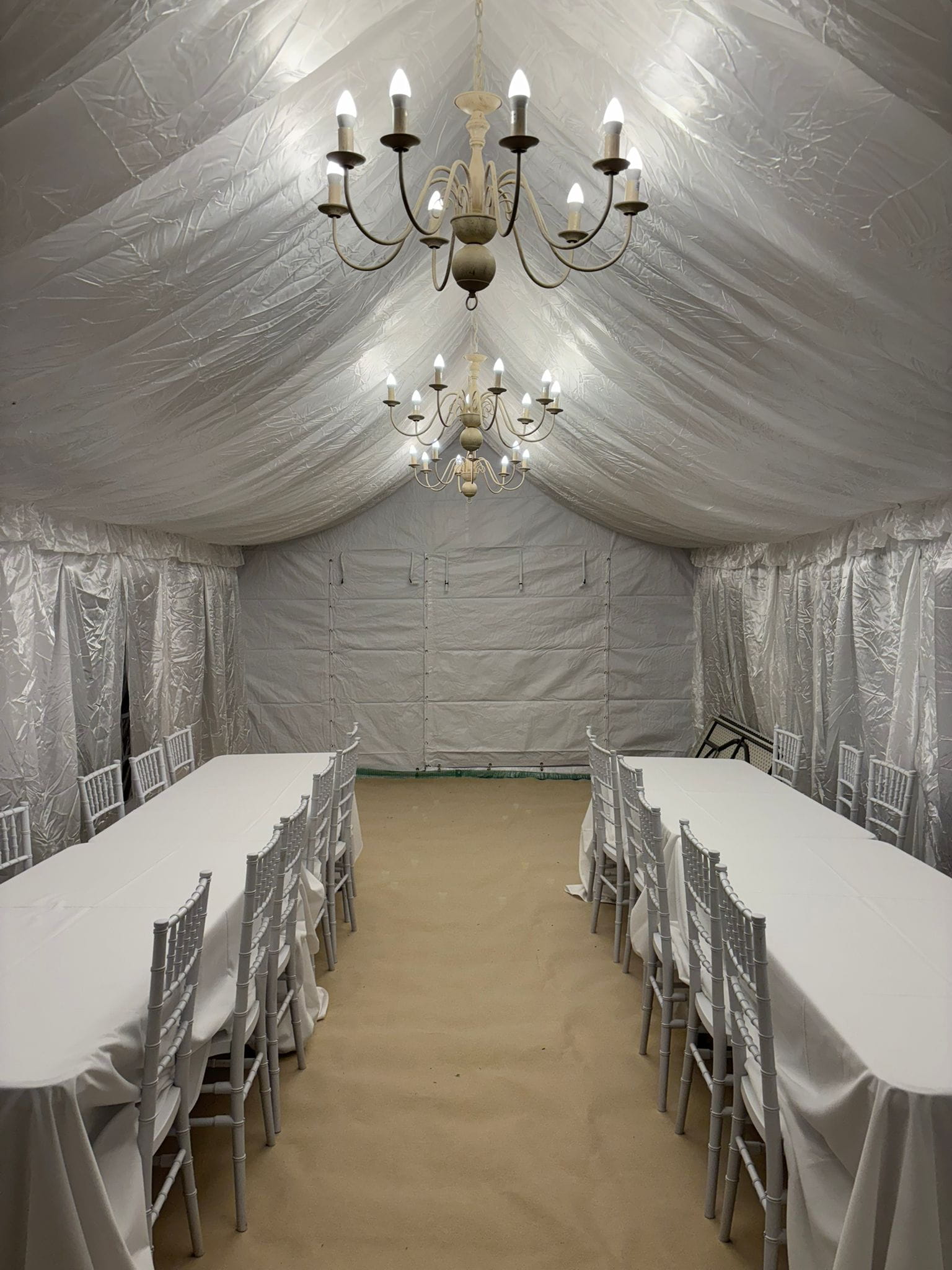 Long-table dining setup with white linens, Chiavari chairs and twin chandeliers in a 4m by 8m marquee